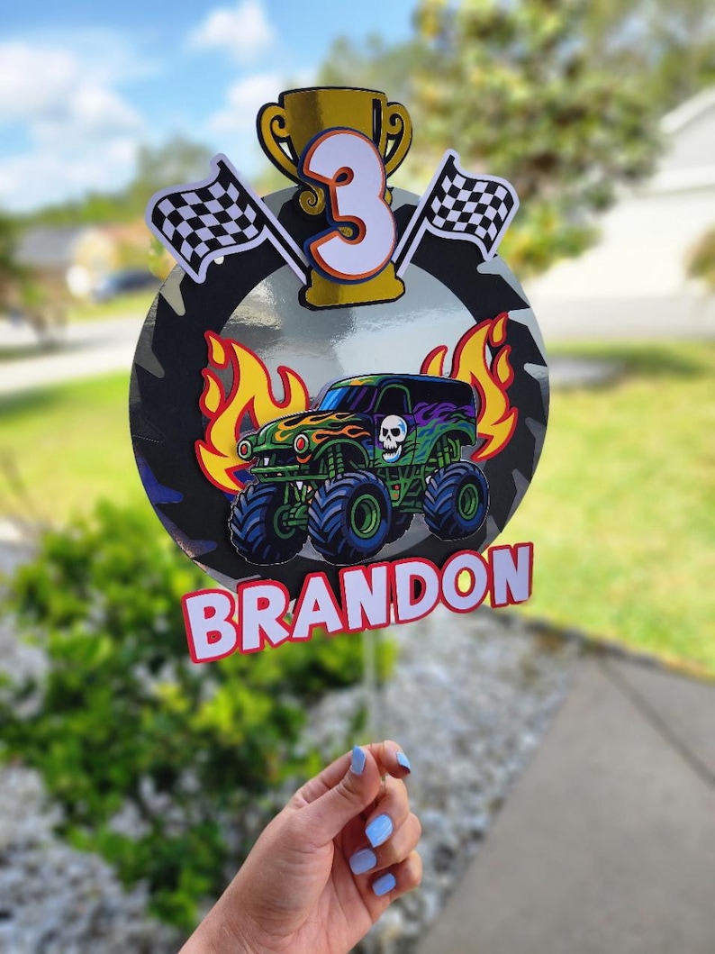 Pode incluir: Decora&ccedil;&atilde;o de anivers&aacute;rio colorida com um monster truck, chamas, uma caveira e o n&uacute;mero 3. Inclui bandeiras quadriculadas, um trof&eacute;u e o nome "BRANDON". O fundo apresenta um padr&atilde;o de camuflagem, ideal para uma festa.