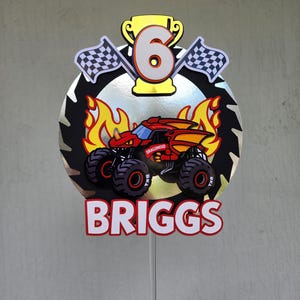 Pode incluir: Topper de bolo de anivers&aacute;rio colorido com um design de monster truck, o nome "BRIGGS" e o n&uacute;mero "6". O topper inclui bandeiras quadriculadas, chamas e um trof&eacute;u, montado em um palito transparente.