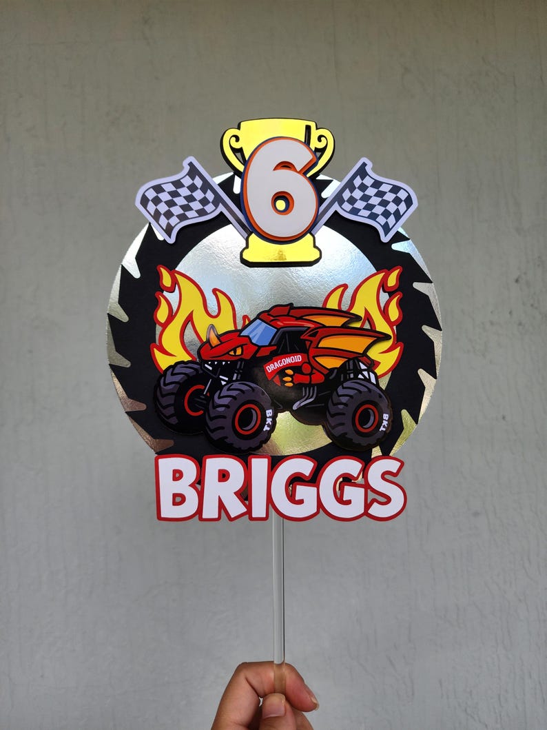 Pode incluir: Topper de bolo de anivers&aacute;rio colorido com um design de monster truck, o nome "BRIGGS" e o n&uacute;mero "6". O topper inclui bandeiras quadriculadas, chamas e um trof&eacute;u, montado em um palito transparente.