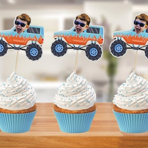 May include: Three cupcakes with blue liners topped with white frosting and blue sprinkles. Each cupcake has a decorative topper featuring a cartoon monster truck with a person's face. The monster trucks are blue and orange.