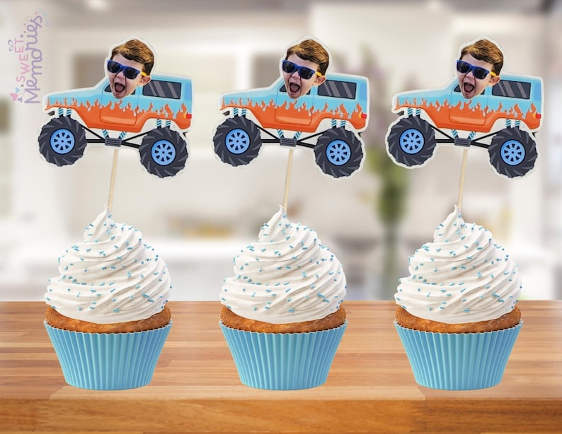 May include: Three cupcakes with blue liners topped with white frosting and blue sprinkles. Each cupcake has a decorative topper featuring a cartoon monster truck with a person's face. The monster trucks are blue and orange.