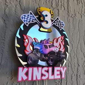 Pode incluir: Topper de bolo de anivers&aacute;rio com um monster truck rosa com chifre de unic&oacute;rnio, chamas e o nome "KINSLEY" em letras rosa. Inclui um trof&eacute;u dourado, o n&uacute;mero 3 e bandeiras quadriculadas.