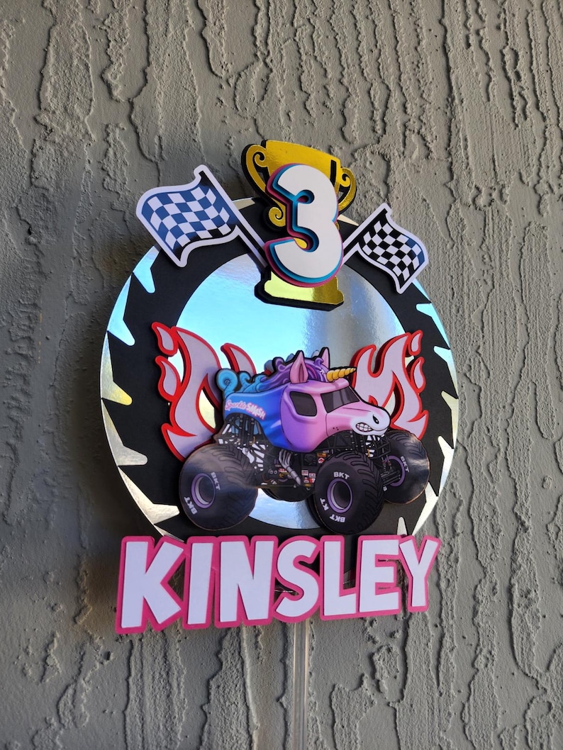 Pode incluir: Topper de bolo de anivers&aacute;rio com um monster truck rosa com chifre de unic&oacute;rnio, chamas e o nome "KINSLEY" em letras rosa. Inclui um trof&eacute;u dourado, o n&uacute;mero 3 e bandeiras quadriculadas.