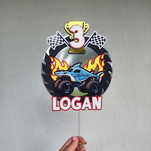Pode incluir: Um topo de bolo colorido com um design de monster truck, um tubar&atilde;o e o texto "MEGALODON". O topo inclui o n&uacute;mero "3", um trof&eacute;u, bandeiras quadriculadas e o nome "LOGAN".