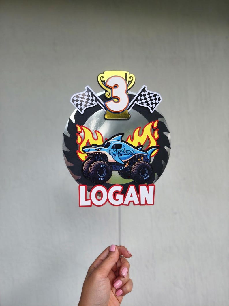 Pode incluir: Um topo de bolo colorido com um design de monster truck, um tubar&atilde;o e o texto "MEGALODON". O topo inclui o n&uacute;mero "3", um trof&eacute;u, bandeiras quadriculadas e o nome "LOGAN".