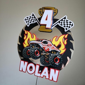 Pode incluir: Topper de bolo de anivers&aacute;rio com um design de monster truck e o nome "Nolan". O topper inclui um trof&eacute;u dourado, o n&uacute;mero "4", bandeiras quadriculadas e chamas. O monster truck &eacute; vermelho e branco.