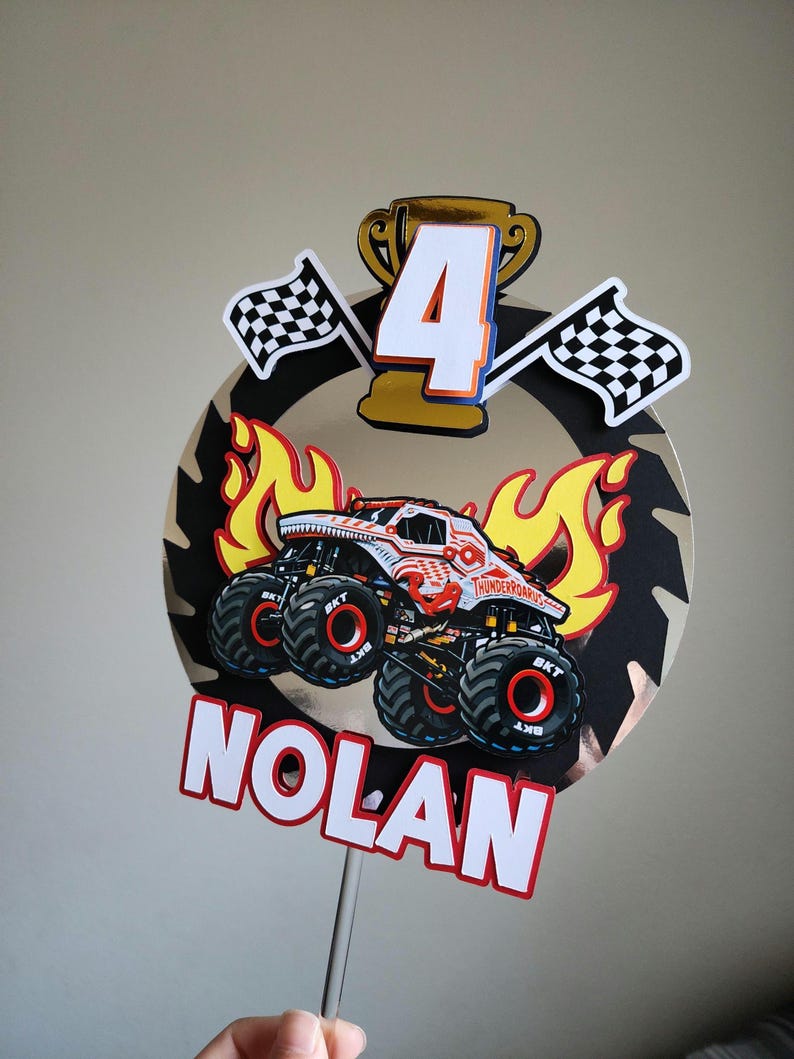 Pode incluir: Topper de bolo de anivers&aacute;rio com um design de monster truck e o nome "Nolan". O topper inclui um trof&eacute;u dourado, o n&uacute;mero "4", bandeiras quadriculadas e chamas. O monster truck &eacute; vermelho e branco.