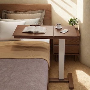 May include: A brown wooden bedside table with a book, coffee cup, and smartphone on top. The table has a white adjustable leg and a brown base. The bed has a brown headboard, white pillows, and a tan blanket.