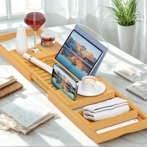 May include: A bamboo bathtub tray holds a tablet, phone, and a cup of coffee. A glass of red wine, a bottle, and a jar are also on the tray. A folded white towel and a book are in the tray.