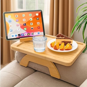 Könnte beinhalten: Ein Sofa-Arm-Tablett aus Bambus mit integriertem Tablethalter. Das Tablett enthält ein Tablet, ein Glas Wasser, einen Teller mit einem Stück Kuchen und Orangenspalten. Das Tablett ist für die Ablage auf einer Sofalehne konzipiert.
