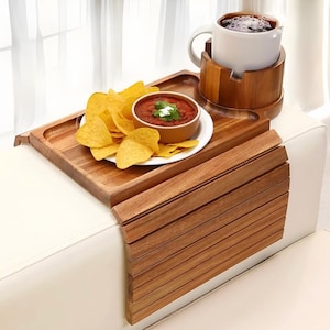 May include: A wooden sofa arm tray with a built-in cup holder. The tray holds a white mug, a small bowl of salsa with a dollop of cream, and a plate of tortilla chips. The tray is made of light brown wood.