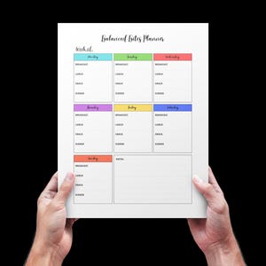 May include: A white paper planner titled "Balanced Bites Planner" is held by two hands against a black background. The planner has a weekly layout with color-coded sections for each day, including spaces for breakfast, lunch, dinner, and snacks.