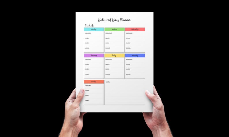 May include: A white paper planner titled "Balanced Bites Planner" is held by two hands against a black background. The planner has a weekly layout with color-coded sections for each day, including spaces for breakfast, lunch, dinner, and snacks.