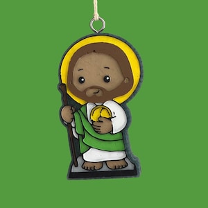 May include: A cartoon-style ornament of Saint Jude, featuring a brown-skinned figure with a brown beard and halo. He wears a white robe, green cloak, and holds a staff and a golden orb. The ornament is suspended by a string against a green background.