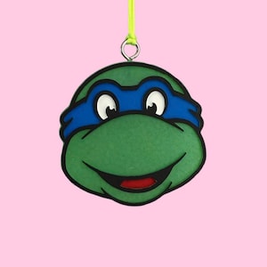 May include: A green turtle character ornament with a blue eye mask, black outlines, and a red mouth. The ornament features a silver metal loop at the top attached to a bright lime green string for hanging, showcasing a flat, graphic design style.