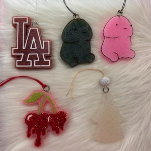 May include: Five hanging ornaments including a maroon and white LA logo, a dark green textured figure, a pink figure with black outlines, a red glittery cherry pair with a green stem, and a translucent white glittery angel-shaped ornament.