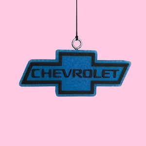 May include: A hanging Chevrolet emblem ornament featuring a bright blue textured surface with a bold black border and the word CHEVROLET embossed in black lettering across the center. The emblem is suspended by a thin black string attached to a metal ring.