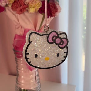 May include: Glittery Hello Kitty-shaped ornament with a pink bow and black outline. The ornament is hanging from a pink string. The background includes a vase with flowers and a pink ribbon.