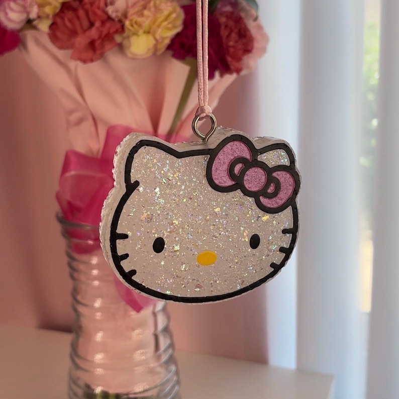 May include: Glittery Hello Kitty-shaped ornament with a pink bow and black outline. The ornament is hanging from a pink string. The background includes a vase with flowers and a pink ribbon.