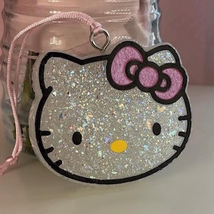 May include: Glittery Hello Kitty-shaped charm with a pink bow and black outline. The charm has a silver glitter interior and a yellow nose. It is attached to a pink string for hanging.