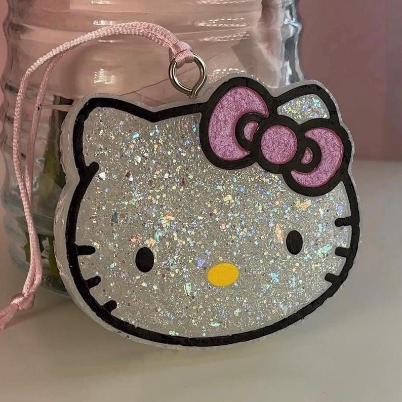 May include: Glittery Hello Kitty-shaped charm with a pink bow and black outline. The charm has a silver glitter interior and a yellow nose. It is attached to a pink string for hanging.