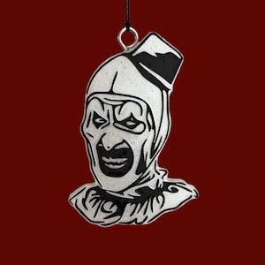 May include: A hanging ornament featuring a stylized black and white illustration of a clown face with a pointed hat. The design includes bold black lines defining the facial features, a ruffled collar, and a small metal loop at the top for hanging.