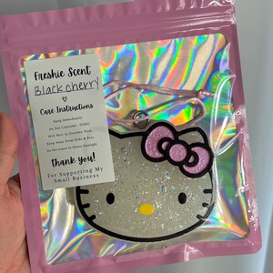 May include: A black cherry scented air freshener in a pink plastic bag. The air freshener is shaped like Hello Kitty with a pink bow. The packaging includes care instructions and a thank you message.