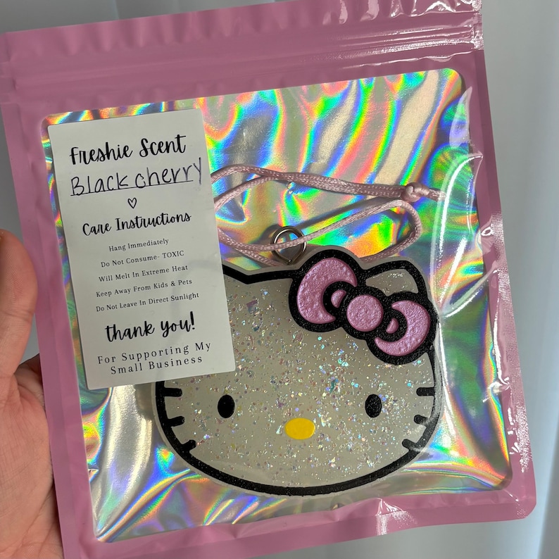 May include: A black cherry scented air freshener in a pink plastic bag. The air freshener is shaped like Hello Kitty with a pink bow. The packaging includes care instructions and a thank you message.