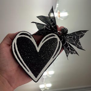 May include: A heart-shaped ornament featuring a black glitter finish with white outlined edges. The ornament is topped with a black ribbon printed with a white spiderweb pattern and a small, dark, textured spherical bead accent attached to the top.