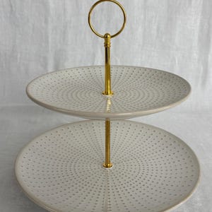 May include: A two-tier serving stand featuring two round, light gray ceramic plates with a radiating pattern of small, dark gray dots. The tiers are connected by a central, polished gold-tone metal rod topped with a circular handle loop.