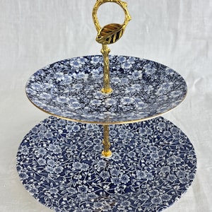 May include: Two-tiered serving stand with blue floral patterned plates and a gold-colored handle. The plates have a dark blue background with white floral designs. The handle is shaped like a branch with a leaf at the top.