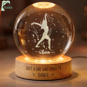 May include: A glowing glass sphere with a dancer silhouette and the name "Olivia" etched on it. The sphere sits on a wooden base with the text "JUST A GIRL WHO LOVES TO DANCE." The base and sphere are illuminated with a warm light.