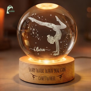 May include: A spherical glass ornament featuring a gymnast design and the name "Olivia". It rests on a wooden base inscribed with "WHY WALK WHEN YOU CAN CARTWHEEL". The base and sphere emit a warm, inviting light.