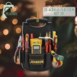 May include: Black tool belt ornament with various tools, including a hammer, screwdrivers, and a multimeter. The ornament has the text "JOHNSON" on the front and "2D ACRYLIC FLAT ITEM NOT 3D" on a tag. The ornament is hanging by a gold string.