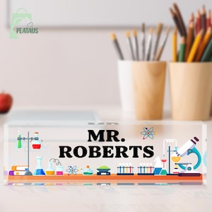 May include: A clear rectangular acrylic desk nameplate featuring the black text MR. ROBERTS centred on the front. The design includes colourful science-themed graphics such as a microscope, various glass beakers, test tubes, and atomic symbols.