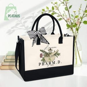 May include: A black and cream tote bag with black handles and a striped bow. The bag features a floral design with the text "Lauren PHARM.D. est 2025". The bag has a black bottom and a detachable shoulder strap.