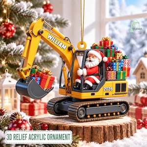 May include: A yellow excavator ornament featuring Santa Claus in the driver seat, the name WILLIAM on the arm, and the year 2026 on the side. The excavator bucket holds a gift, and a stack of colorful wrapped presents sits on the back of the vehicle.
