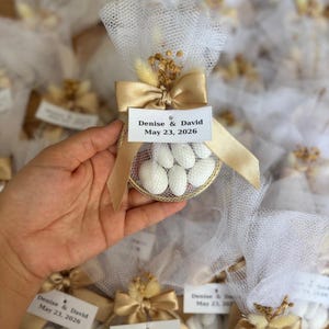 May include: A gold-coloured favour bag with white tulle, a gold ribbon, and a tag that reads "Denise & David May 23, 2026." The bag contains white candies. Other similar bags are in the background.