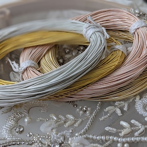 May include: Three spools of metallic wire, one silver, one gold, and one rose gold, are stacked on top of each other. The spools are on a white background with a beaded design.
