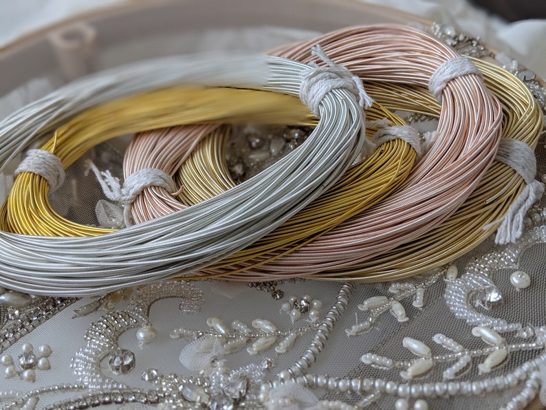May include: Three spools of metallic wire, one silver, one gold, and one rose gold, are stacked on top of each other. The spools are on a white background with a beaded design.