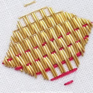 May include: Close-up of a gold metallic thread embroidery design on white fabric. The design is a woven pattern with a pink thread outlining the edges.