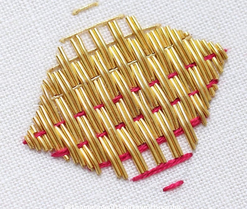May include: Close-up of a gold metallic thread embroidery design on white fabric. The design is a woven pattern with a pink thread outlining the edges.