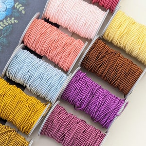 May include: Assortment of colourful elastic cord spools in various colours, including light blue, peach, yellow, brown, purple, and gold.