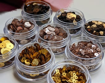 Op de afbeelding: Assortiment van kleine potjes gevuld met verschillende kleuren ronde pailletten. De kleuren omvatten goud, brons, bruin en zwart.