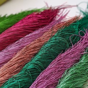 May include: A collection of colorful, long, thin strands of yarn in shades of green, pink, red, and brown. The strands are arranged in a fan shape, with the tips of the strands pointing towards the right side of the image.