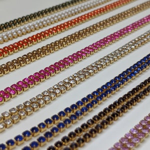 May include: A close-up of ten rows of gold chains with different colored square rhinestones. The chains are arranged in a staggered pattern, with each row slightly offset from the one above it. The colors of the rhinestones are: black, blue, clear, pink, red, and brown.