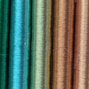Op de afbeelding: Een reeks verticale klossen garen gerangschikt in een kleurverloop. De kleuren gaan over van diep smaragdgroen en helder turkoois naar licht olijfgroen, gevolgd door warm koper, brons en uiteindelijk een goudgele tint.