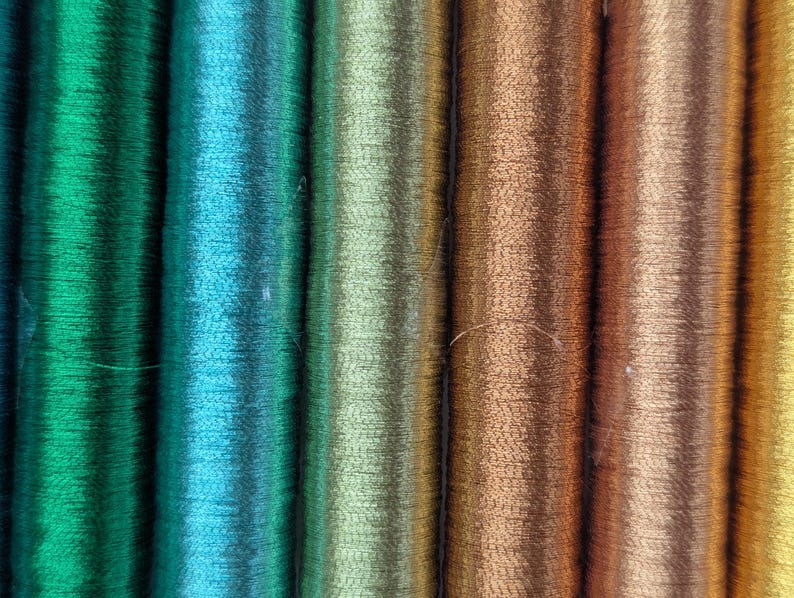 Op de afbeelding: Een reeks verticale klossen garen gerangschikt in een kleurverloop. De kleuren gaan over van diep smaragdgroen en helder turkoois naar licht olijfgroen, gevolgd door warm koper, brons en uiteindelijk een goudgele tint.