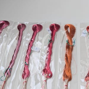 Op de afbeelding: Zes individuele strengen borduurgaren, elk verzegeld in een doorzichtige plastic hoes. De kleuren vari&euml;ren van diep magenta en verschillende roze tinten tot een fel oranje en een rood-oranje tint. Elke streng is voorzien van een wit papieren label.
