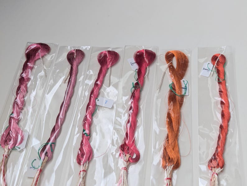 Op de afbeelding: Zes individuele strengen borduurgaren, elk verzegeld in een doorzichtige plastic hoes. De kleuren vari&euml;ren van diep magenta en verschillende roze tinten tot een fel oranje en een rood-oranje tint. Elke streng is voorzien van een wit papieren label.
